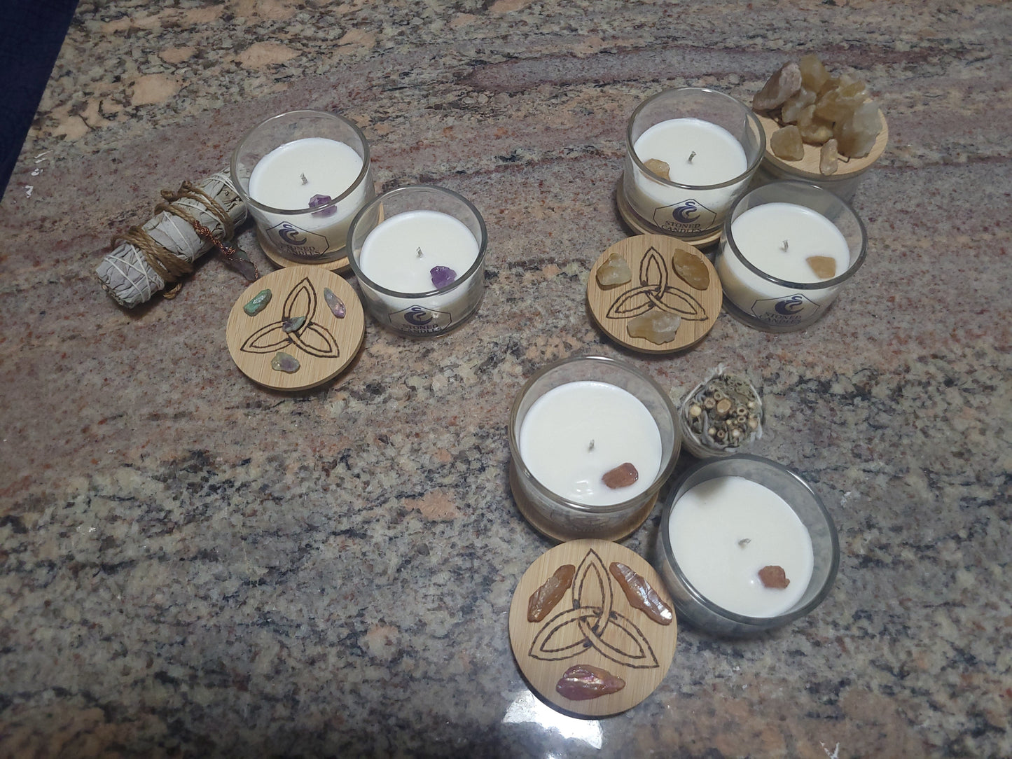 Handcrafted Awen Aboros gemstone candles in glass vessels with engraved triquetra lids, embedded crystals, and sage bundles — custom ritual candle collection
