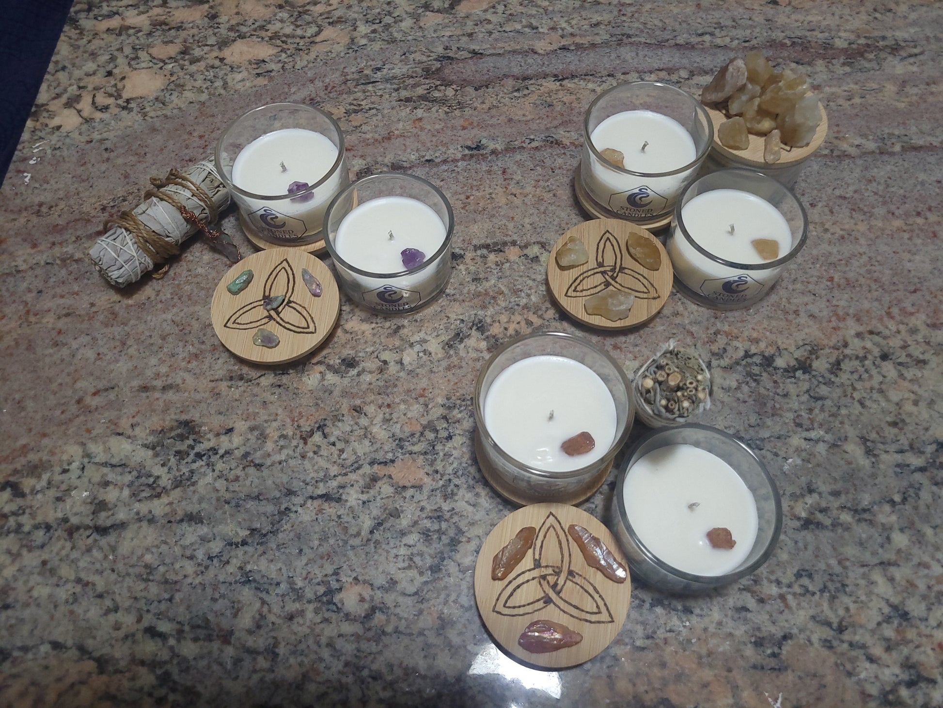 Handcrafted Awen Aboros gemstone candles in glass vessels with engraved triquetra lids, embedded crystals, and sage bundles — custom ritual candle collection