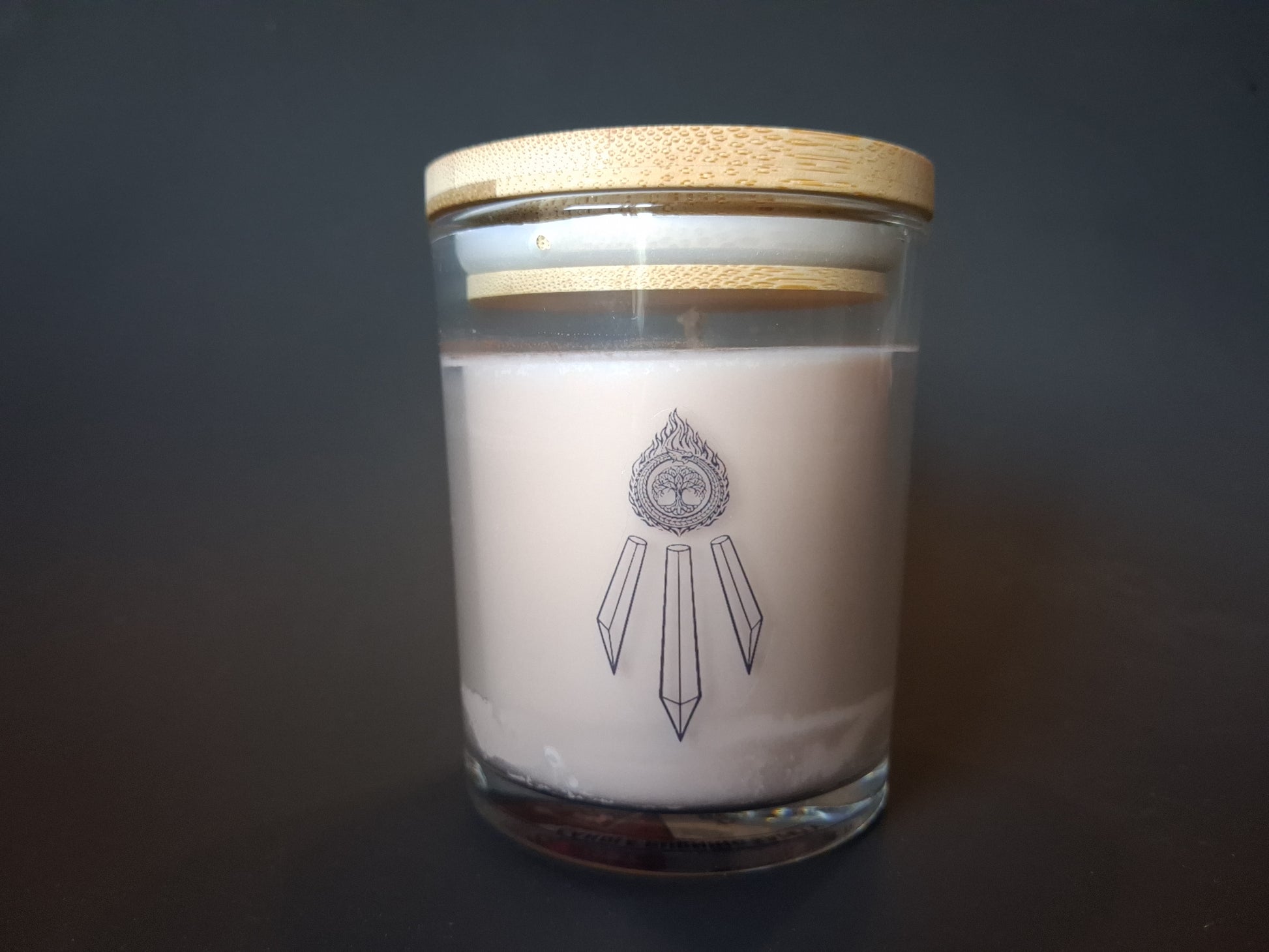 The Knowing candle in a glass jar with bamboo lid, cream wax with Awen Aboros crystal logo etched on glass, dark background