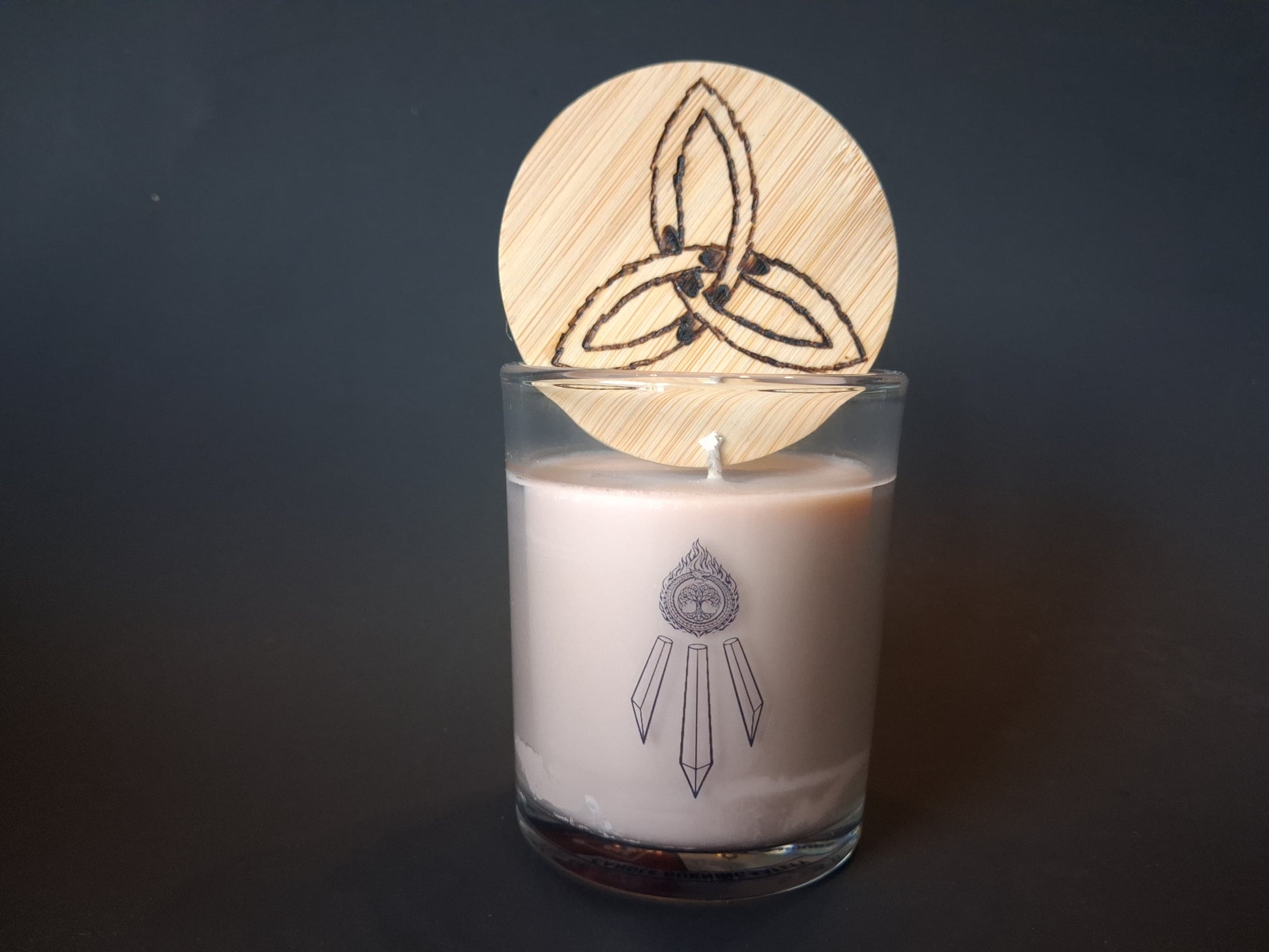 The Knowing candle with engraved triquetra wooden lid resting on top, cream wax in clear glass with Awen Aboros logo