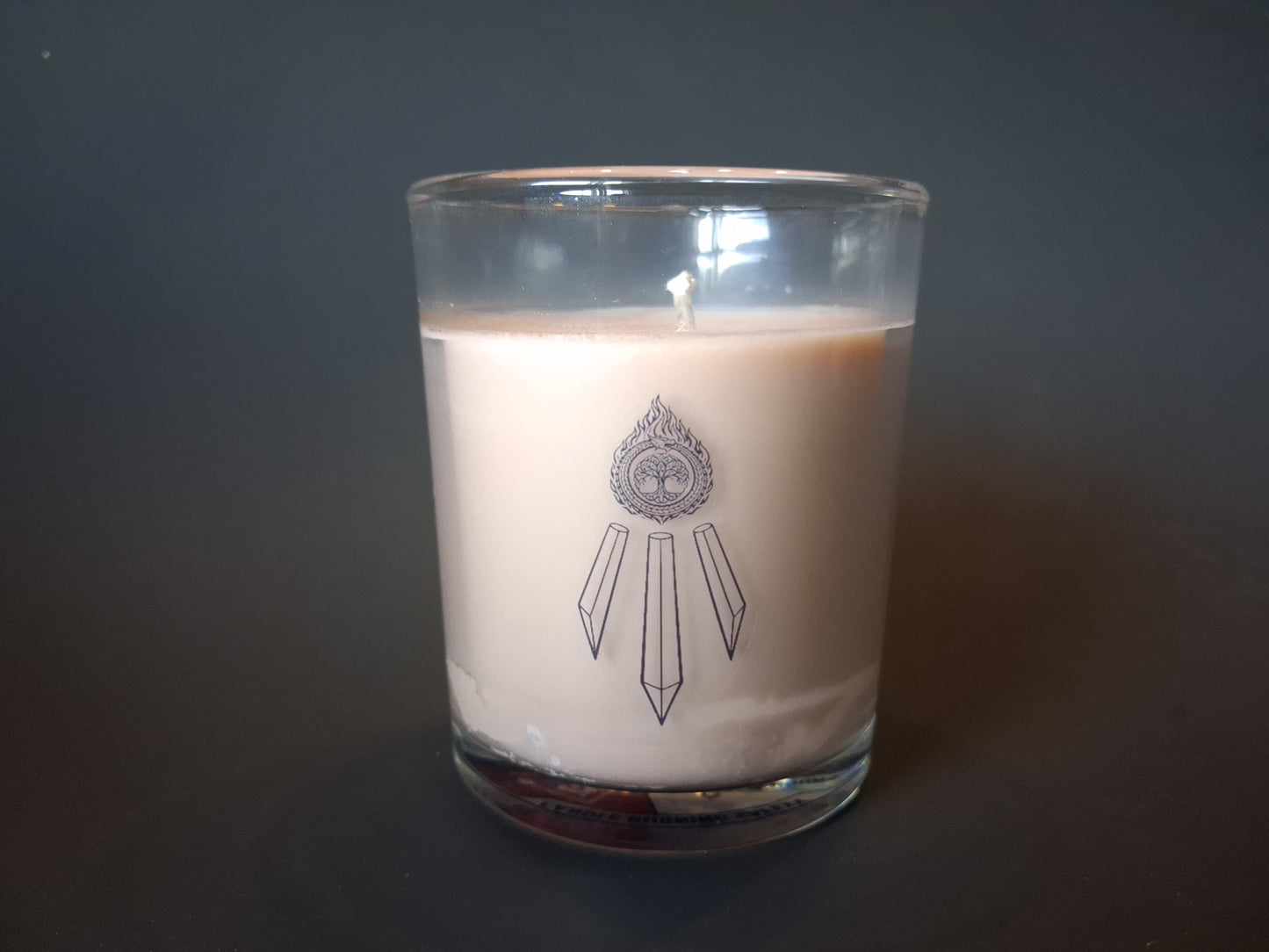 The Knowing candle without lid, cream wax with visible wick and Awen Aboros crystal logo etched on clear glass, dark background