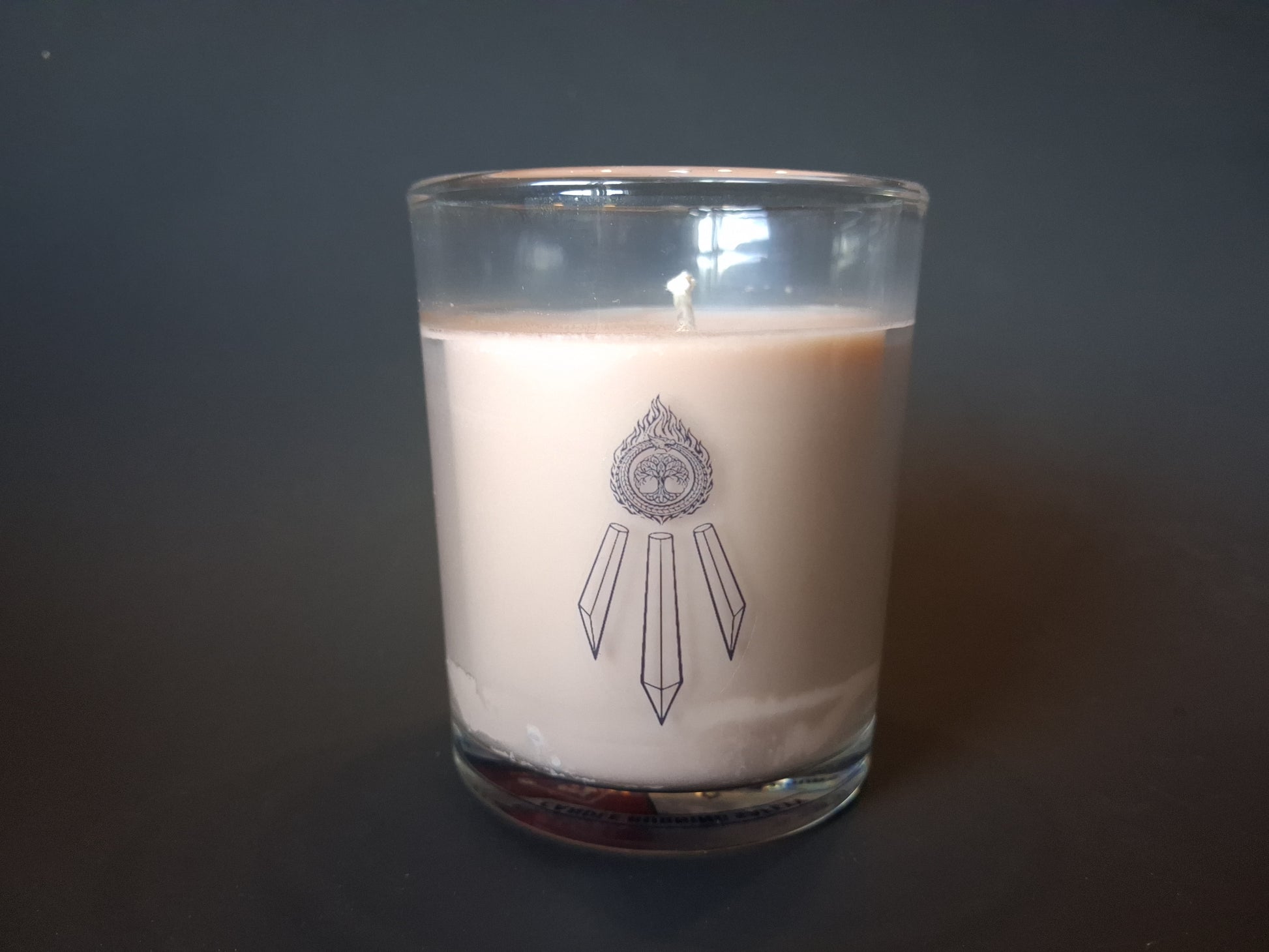 The Knowing candle without lid, cream wax with visible wick and Awen Aboros crystal logo etched on clear glass, dark background