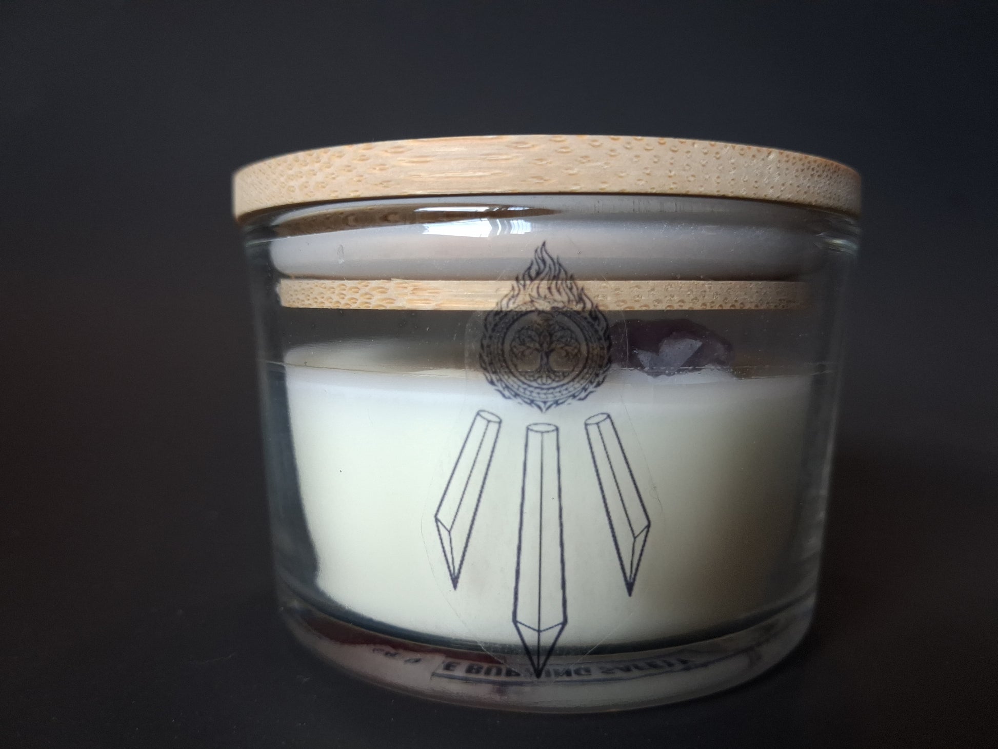 Amethyst Intentions candle with plain bamboo lid on wide-mouth clear glass jar, raw amethyst gemstone visible through glass, cream wax with Awen Aboros crystal logo, dark background