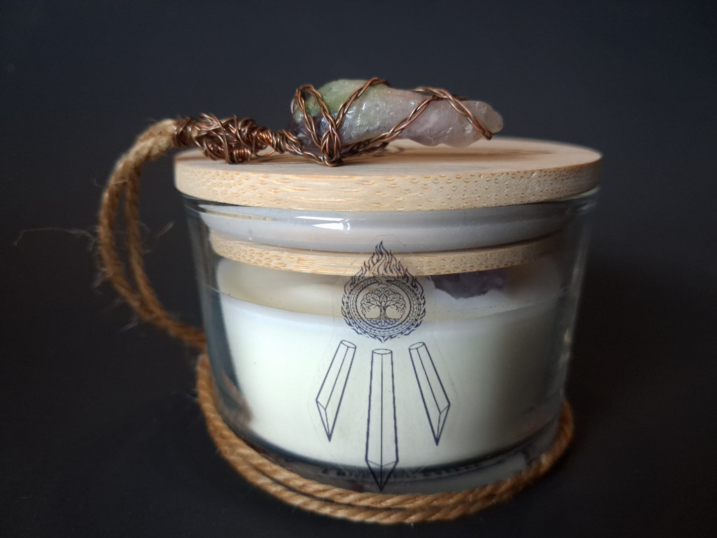Amethyst Intentions candle with bamboo lid topped with wire-wrapped raw crystal pendant on jute cord, cream wax with Awen Aboros logo, dark background