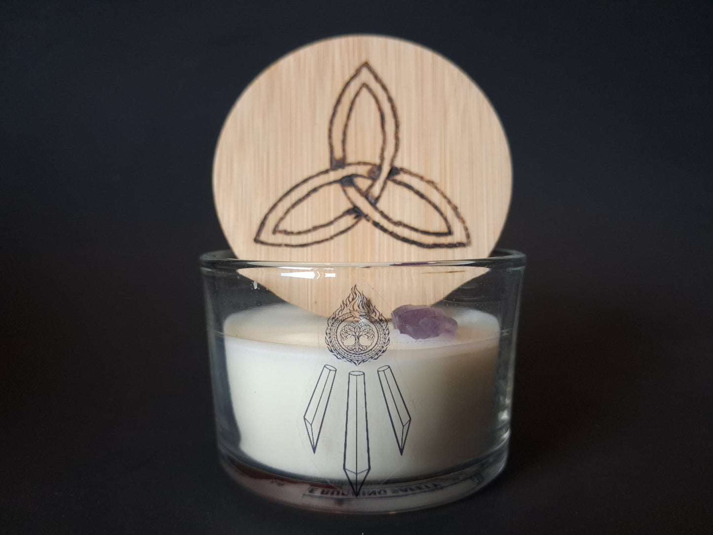 Amethyst Intentions candle with engraved triquetra wooden lid propped open, raw amethyst gemstone visible atop cream wax, Awen Aboros logo on clear glass, dark background