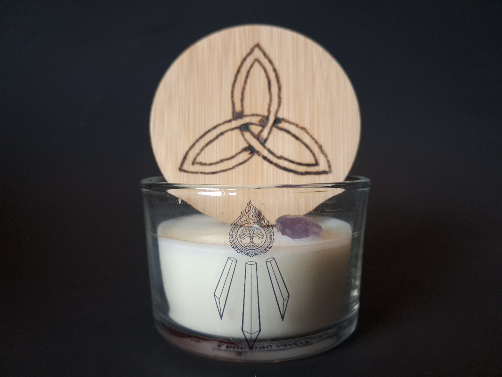 Amethyst Intentions candle with engraved triquetra wooden lid propped open, raw amethyst gemstone visible atop cream wax, Awen Aboros logo on clear glass, dark background