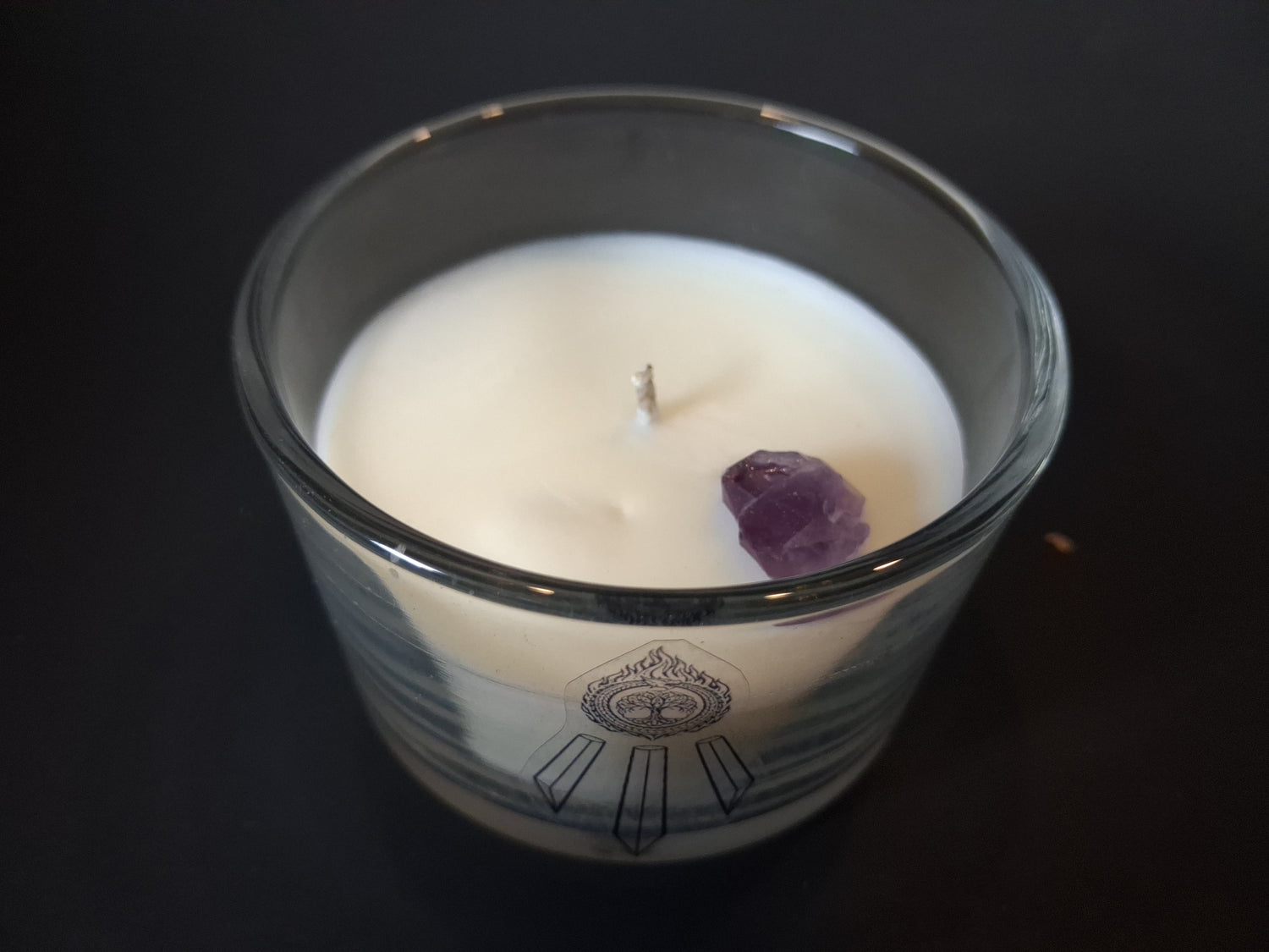 Amethyst Intentions candle top-down view showing raw amethyst gemstone and wick set in cream wax, Awen Aboros logo etched on clear glass, dark background