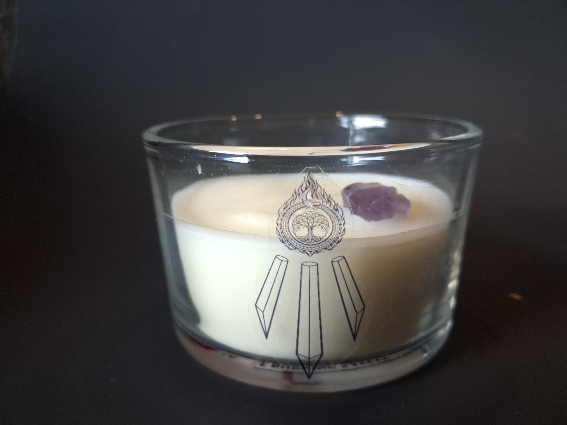 Amethyst Intentions candle without lid, raw amethyst gemstone resting atop cream wax, Awen Aboros crystal and tree logo etched on wide-mouth clear glass, dark background