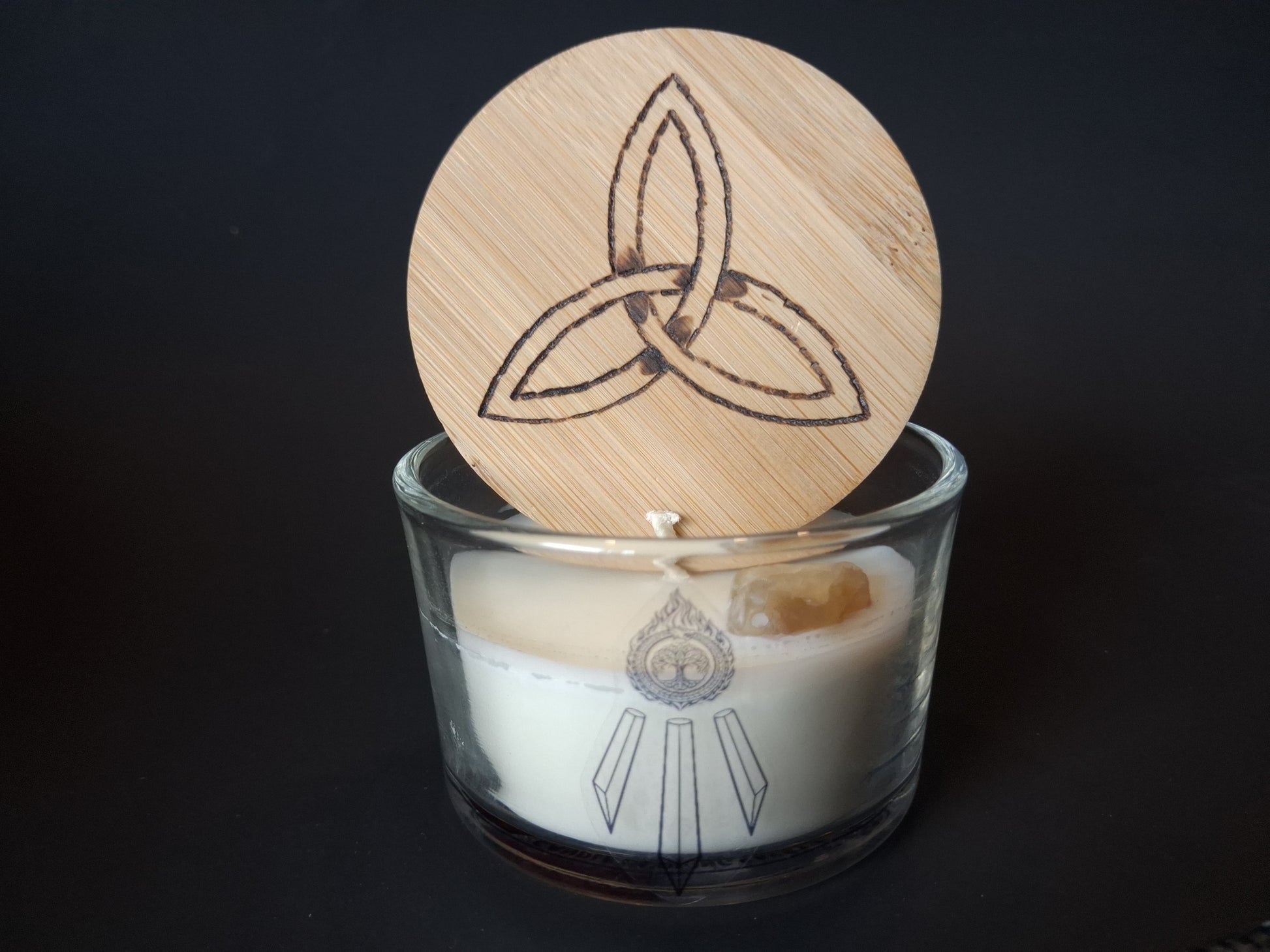 Citrine Intentions candle with engraved triquetra wooden lid propped open, raw citrine gemstone visible in cream wax, Awen Aboros logo on clear glass, dark background