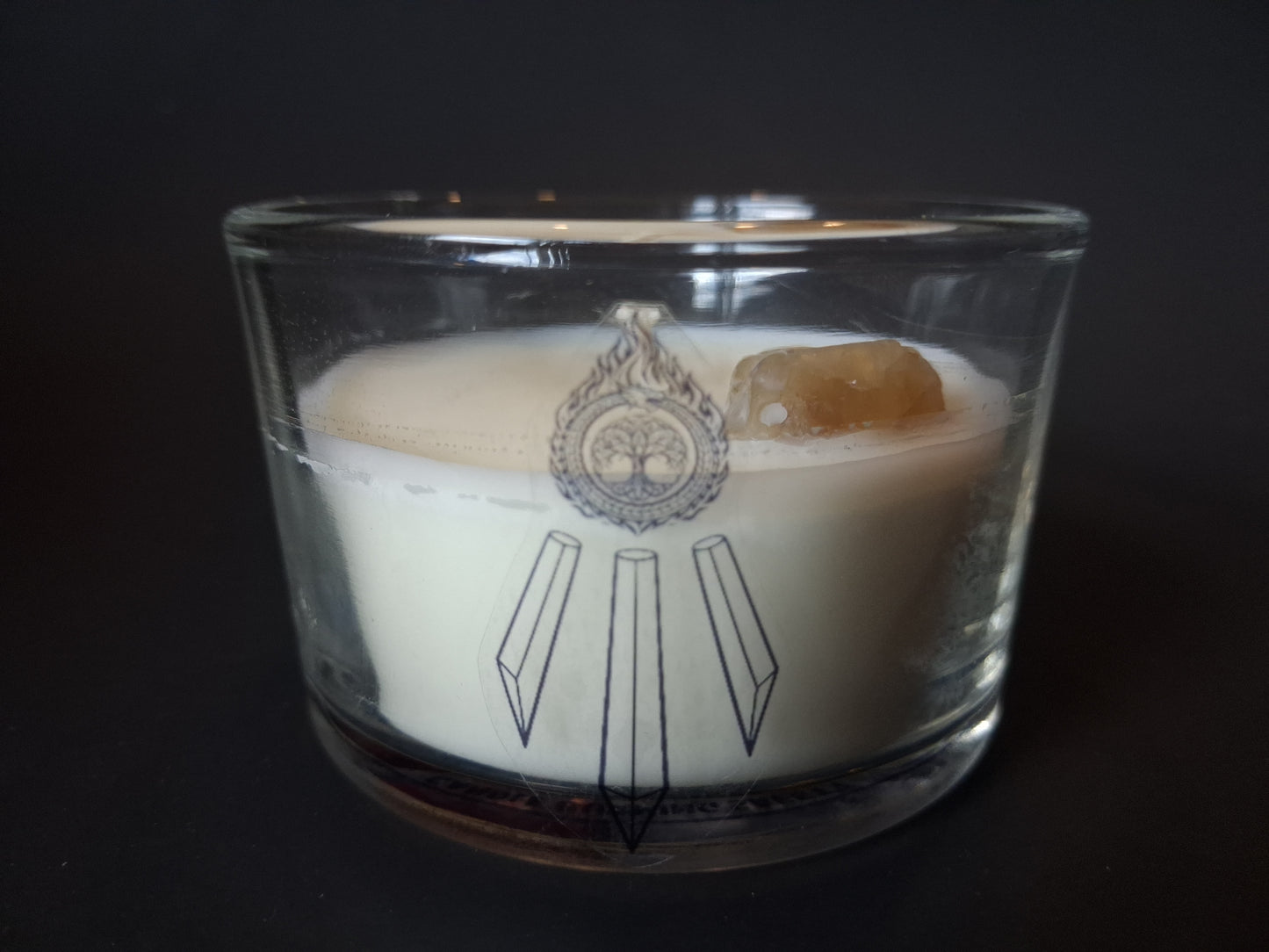 Citrine Intentions candle without lid, raw citrine gemstone visible in cream wax, Awen Aboros crystal and tree logo etched on wide-mouth clear glass, dark background