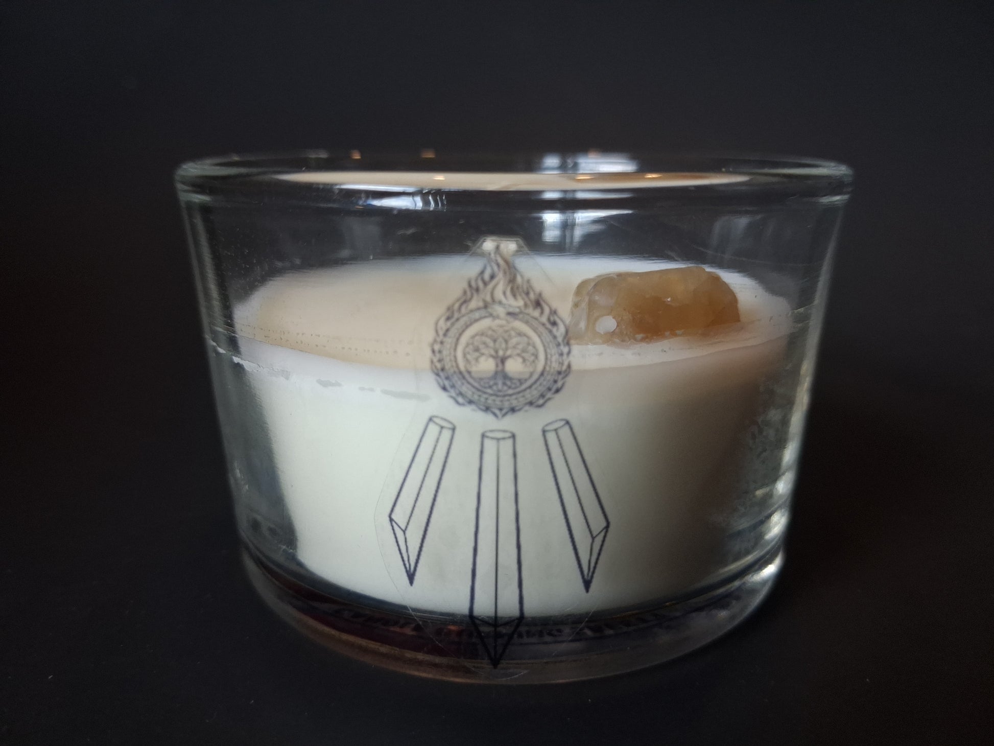 Citrine Intentions candle without lid, raw citrine gemstone visible in cream wax, Awen Aboros crystal and tree logo etched on wide-mouth clear glass, dark background