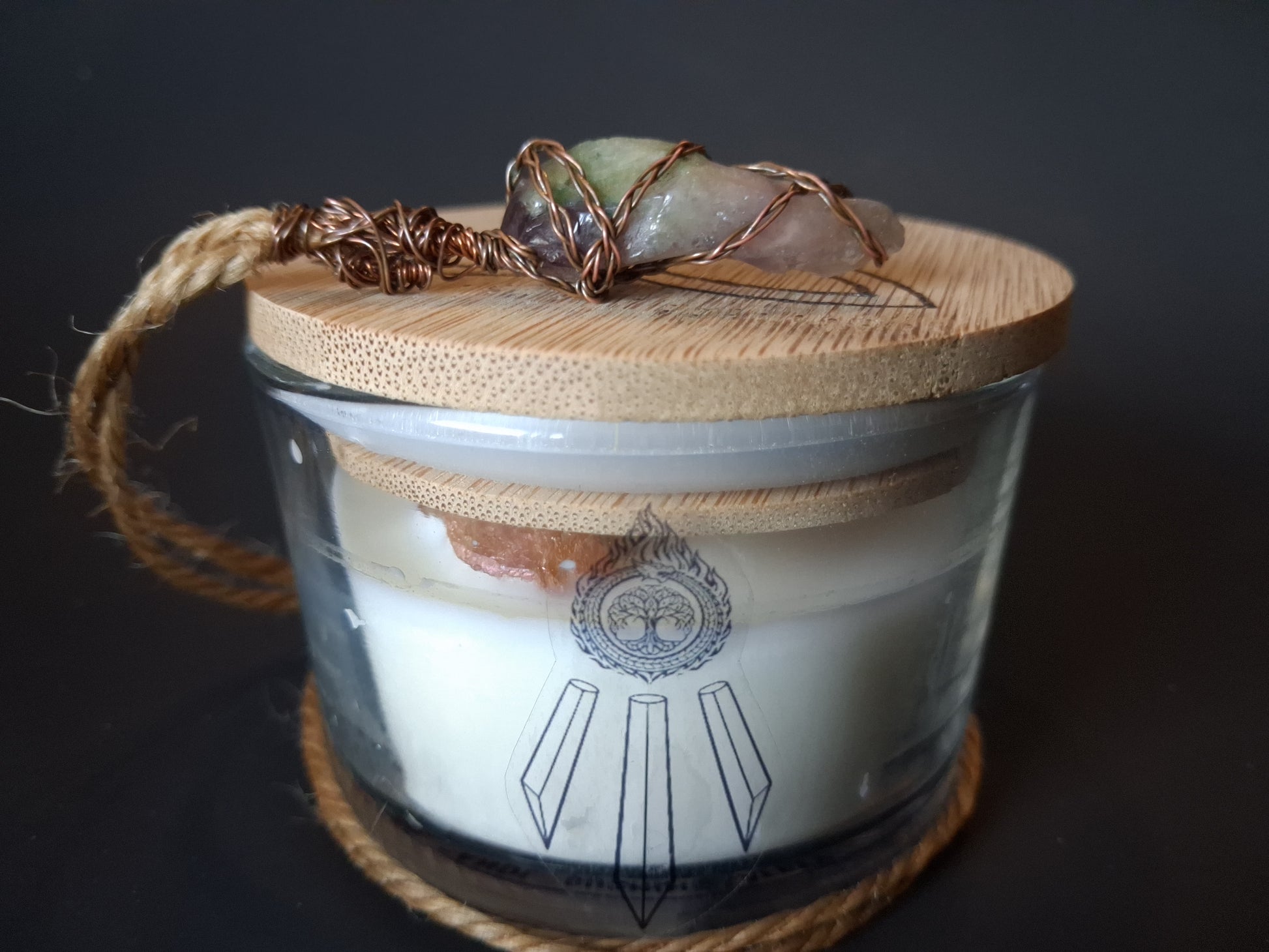 Champagne Aura Intentions candle with bamboo lid topped with wire-wrapped multi-stone crystal pendant on jute cord, cream wax with Awen Aboros logo, dark background