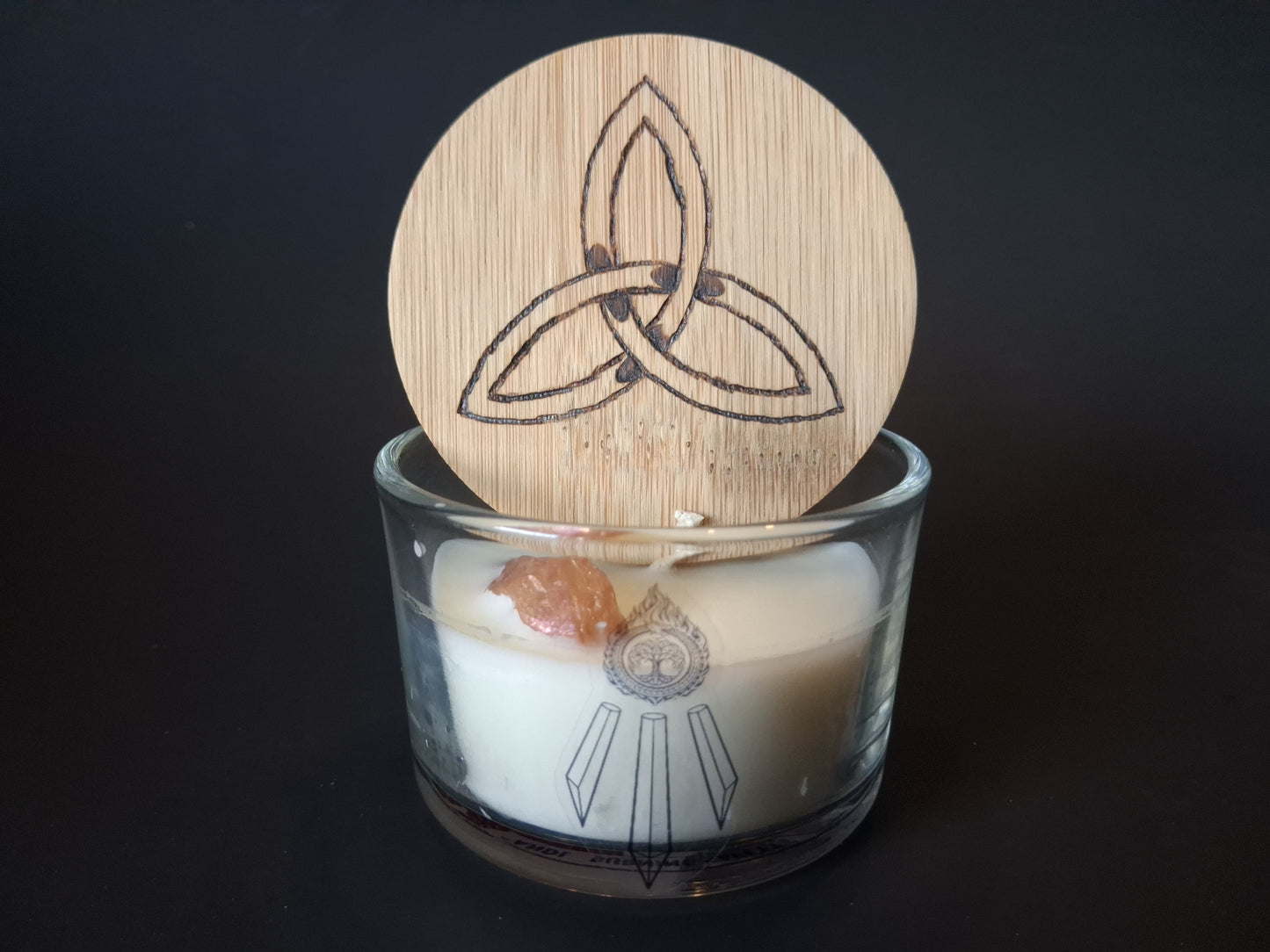 Champagne Aura Intentions candle with engraved triquetra wooden lid propped open, raw champagne aura quartz gemstone embedded in cream wax, Awen Aboros logo on clear glass, dark background