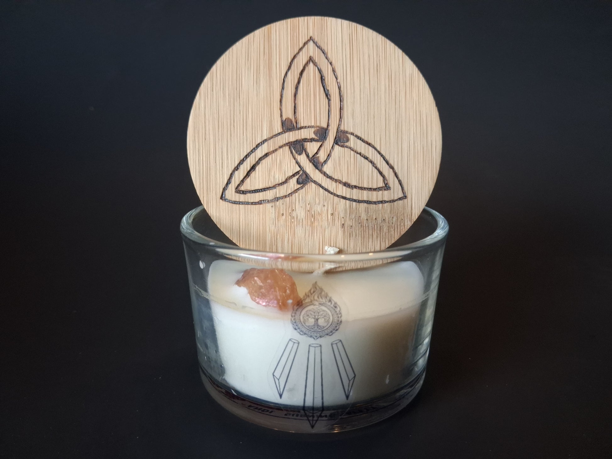 Champagne Aura Intentions candle with engraved triquetra wooden lid propped open, raw champagne aura quartz gemstone embedded in cream wax, Awen Aboros logo on clear glass, dark background