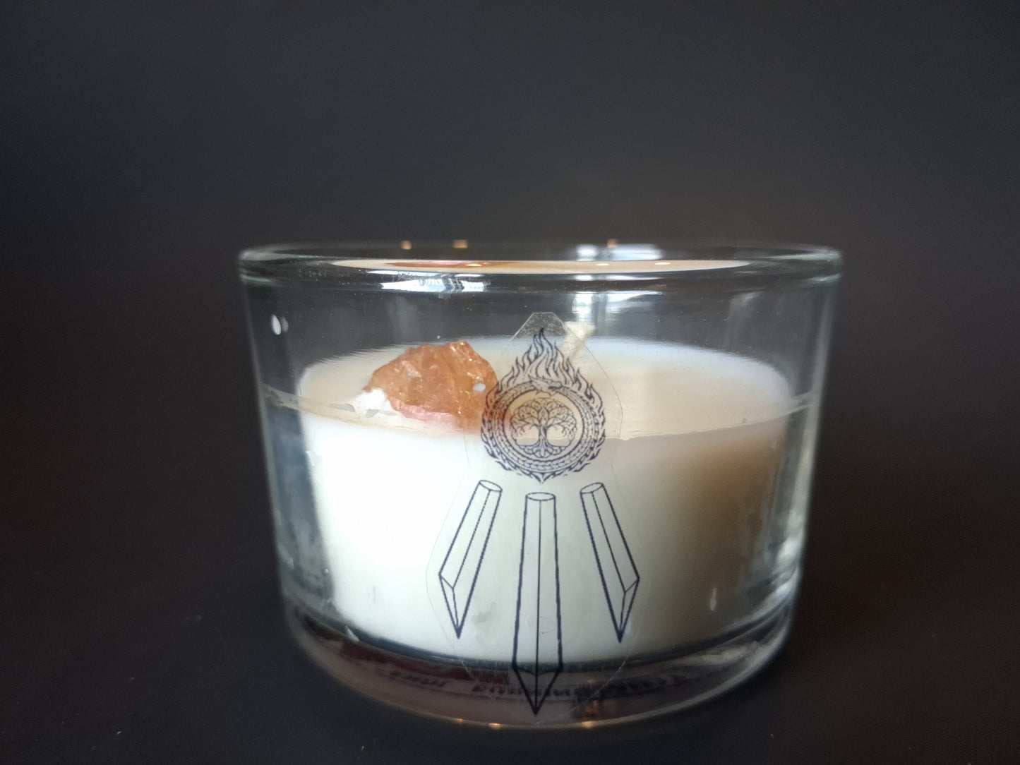 Champagne Aura Intentions candle without lid, raw champagne aura quartz gemstone visible through clear glass jar, cream wax with Awen Aboros crystal logo, dark background