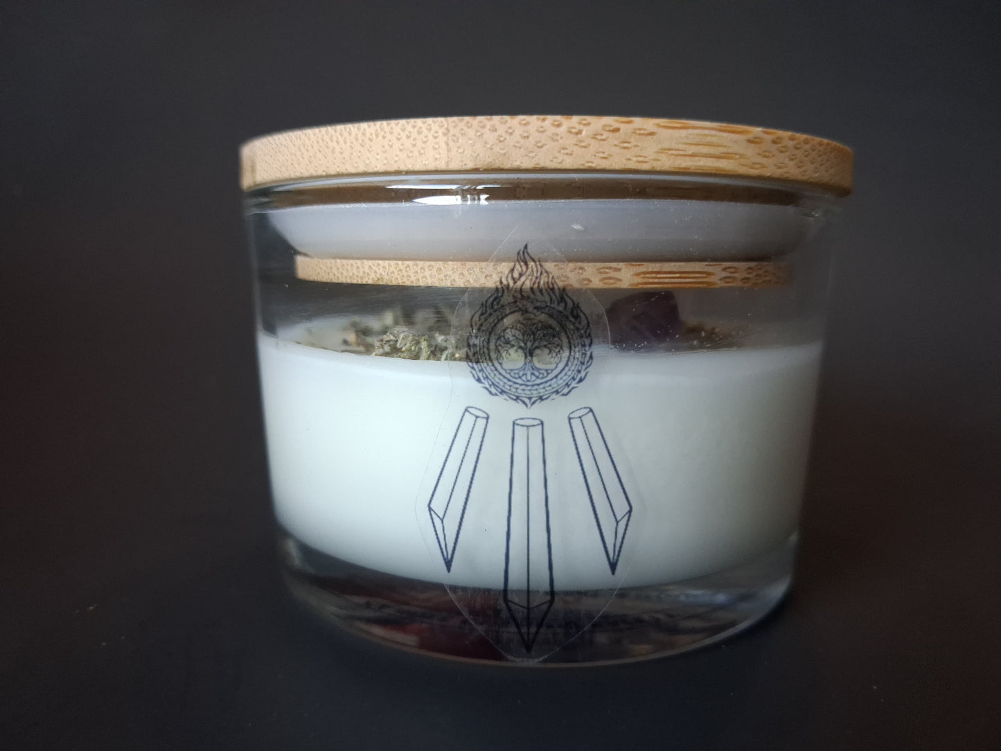 Soft Ground candle with plain bamboo lid on clear glass jar, dried white sage herbs and amethyst crystal visible through glass, Awen Aboros tree and crystal logo, dark background