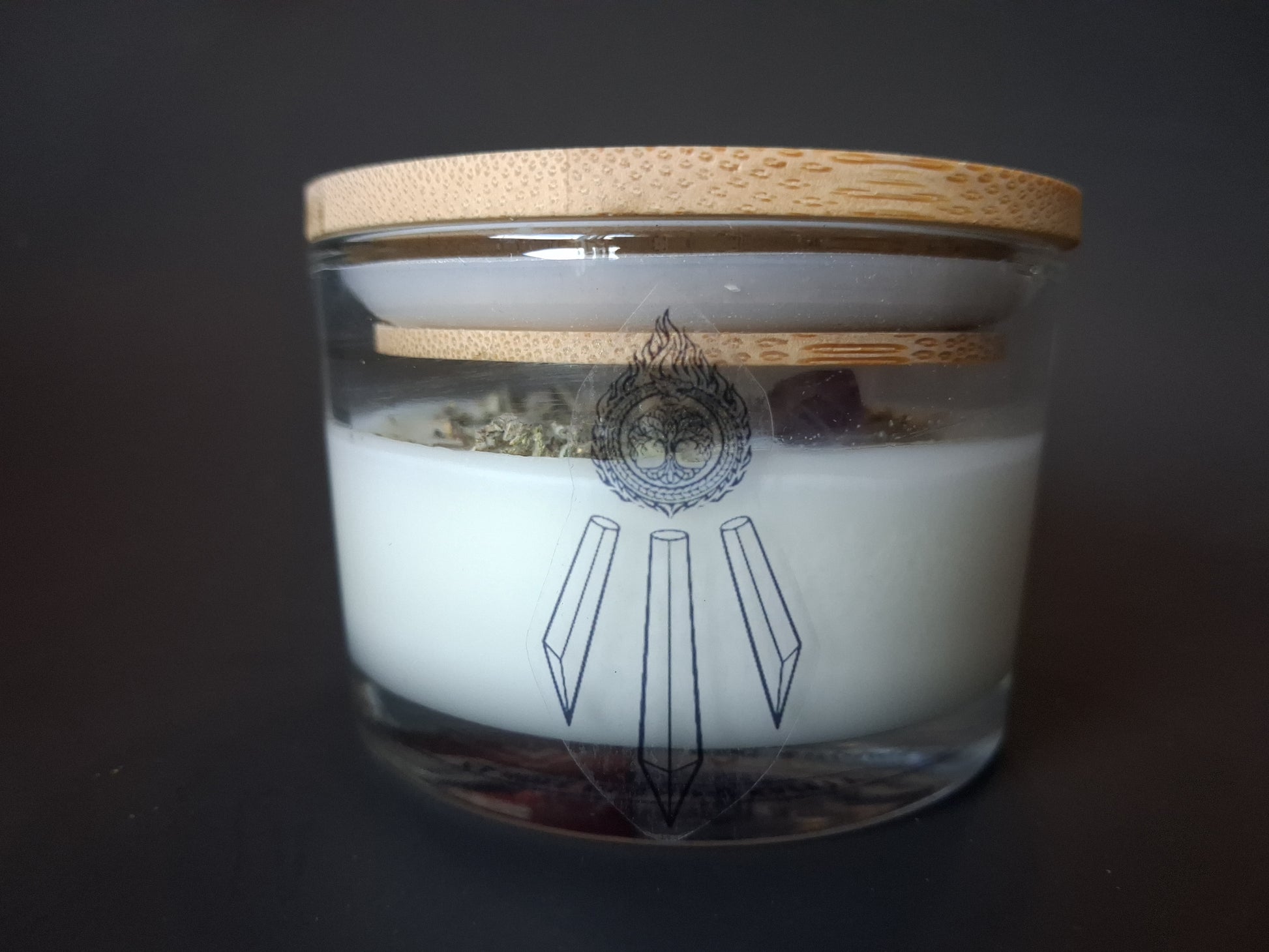 Soft Ground candle with plain bamboo lid on clear glass jar, dried white sage herbs and amethyst crystal visible through glass, Awen Aboros tree and crystal logo, dark background