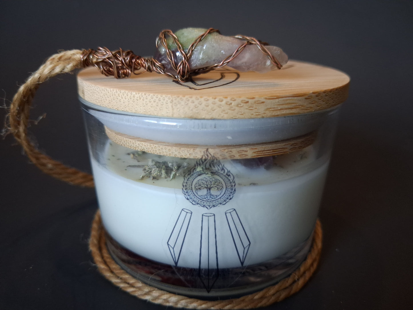Soft Ground candle with bamboo lid topped with wire-wrapped amethyst pendant on jute cord, cream wax with Awen Aboros tree logo on clear glass, dark background