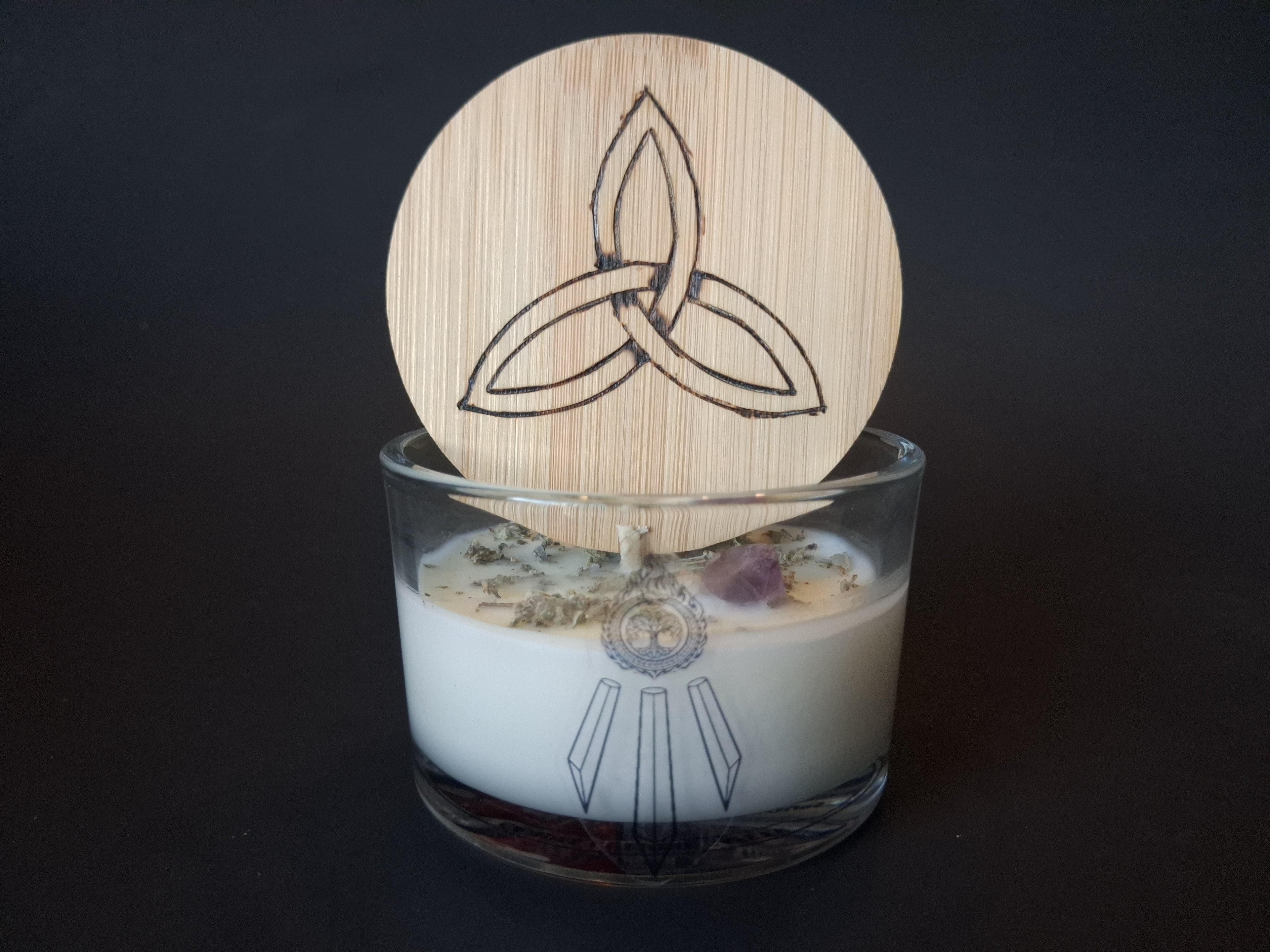 Soft Ground candle with engraved triquetra wooden lid propped open, dried white sage and raw amethyst gemstone visible in cream wax, Awen Aboros logo on clear glass, dark background