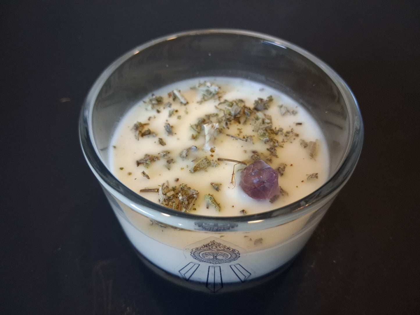 Soft Ground candle close-up top-down view showing dried white sage and raw amethyst gemstone set in cream wax, wick centered, Awen Aboros logo on wide-mouth clear glass, dark background