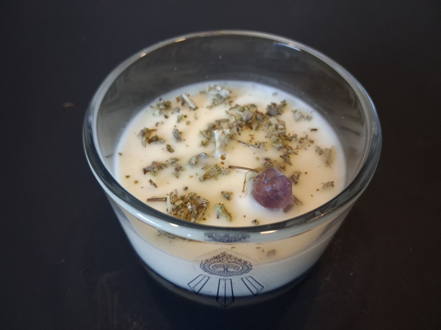 Soft Ground candle close-up top-down view showing dried white sage and raw amethyst gemstone set in cream wax, wick centered, Awen Aboros logo on wide-mouth clear glass, dark background
