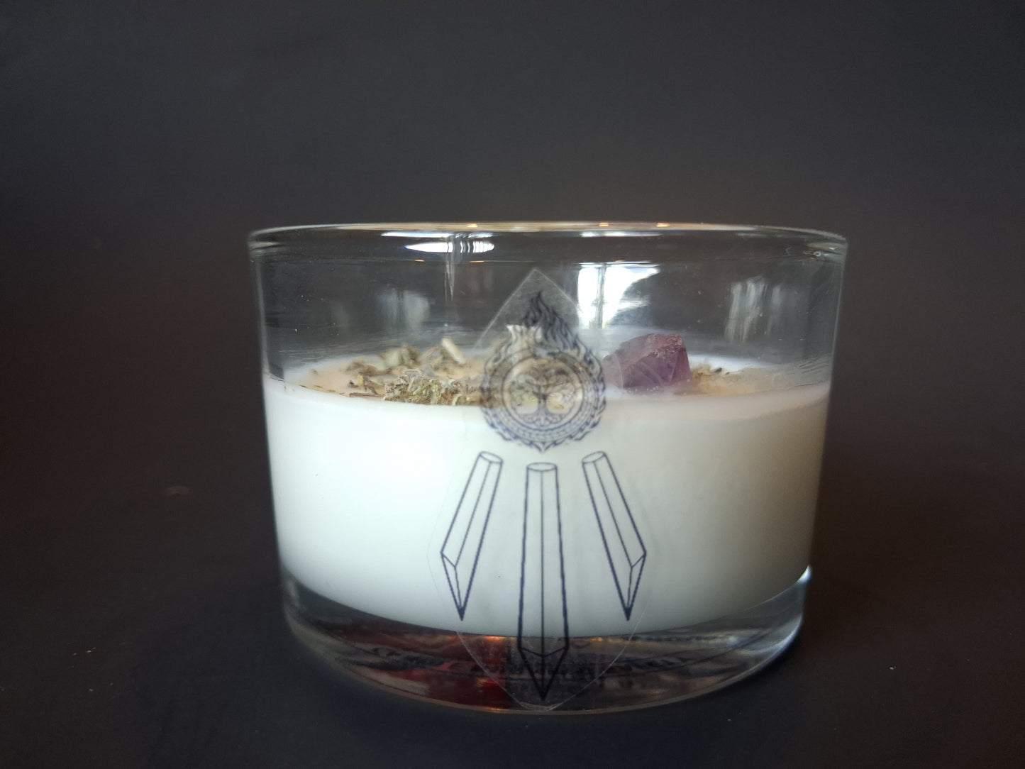 Soft Ground candle top-down view showing dried lavender herbs, white sage, raw amethyst gemstone and wick set in cream wax, Awen Aboros logo on clear glass, dark background